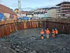 Anleggsarbeidere i oransje verneklær står i en dyp byggegrop omgitt av spuntvegger i bymiljø.