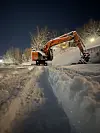 Oransje gravemaskin rydder snø langs en snødekt vei en mørk vinterkveld.