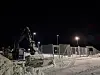 Gravemaskin og gjerder ved snødekt grenseområde en mørk vinterkveld.
