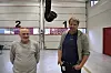 To personer står i en lys verkstedhall foran store porter merket med tall.