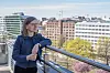 En kvinne står ved et rekkverk på en takterrasse med bybygg og trær i bakgrunnen.