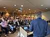 Et fullt auditorium med mange sittende deltakere og en taler sett bakfra ved en talerstol.