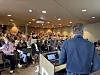 Et fullt auditorium med mange sittende deltakere og en taler sett bakfra ved en talerstol.