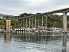 En høy bru strekker seg over vannet med støttepilarer, bygg og båter i forgrunnen.
