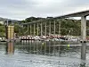 En høy bru strekker seg over vannet med støttepilarer, bygg og båter i forgrunnen.