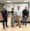 Fire personer står i et møterom foran et lerret, med en klokke på veggen og bord langs bakveggen.