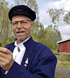 Mann med caps og briller står utendørs foran trær og en rød bygning.