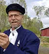 Mann med caps og briller står utendørs foran trær og en rød bygning.