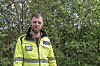 En mann i gul refleksjakke står foran grønne trær og busker.