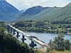 En bru under bygging går over en fjord med fjell i bakgrunnen og anleggsvei langs vannet.