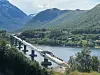 En bru under bygging går over en fjord med fjell i bakgrunnen og anleggsvei langs vannet.