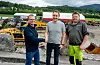 Tre menn står på et anleggsområde med gravemaskiner og lastebiler i bakgrunnen.