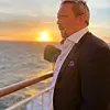En mann i mørk dress står ved rekverket på et dekk mens solen går ned over havet.