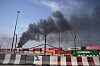 Mørk røyk stiger over en havn med containere, gjerder og industribygg.
