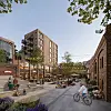 Illustrasjon av et urbant område med boligbygg, mennesker, syklist og uteservering ved en åpen plass.