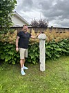 En mann står på gresset ved siden av en høy, lys betongsøyle foran en hekk og et hus.
