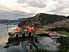 Stort offshoreanlegg ved kai med kraner, røde fartøy og fjell i bakgrunnen.