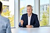 En mann i mørk dress sitter ved et bord i et lyst møterom og snakker med en person i forgrunnen.