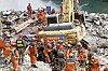 Redningsarbeidere i oransje og røde hjelmer søker i ruiner ved en gravemaskin nær vannet.