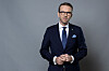 En mann i mørk dress og briller står mot grå bakgrunn med hendene samlet foran seg.