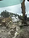 Gravemaskin arbeider i et nedbrent område med røyk, ruiner og brannskader i terrenget.