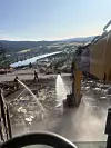Utsikt over et brannområde med utbrente ruiner, gravemaskin og vannstråler i arbeid.