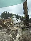 En gravemaskin arbeider blant ruiner og brannrester mens damp stiger opp i et nedbrent område.