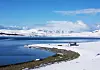 Snødekt kystlinje ved en delvis islagt fjord med en liten bygning og fjell i bakgrunnen.