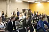 Studenter sitter og står i en lys sal med stoler, mens folk samles foran en murvegg og skjermer.