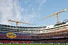 Fotballstadion med åpne tribuner og to gule kraner mot en blå himmel.