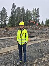 Person i gul hjelm og jakke står på en steinete anleggsplass med gravemaskin i bakgrunnen.