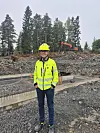 Person i gul hjelm og jakke står på en steinete anleggsplass med gravemaskin i bakgrunnen.