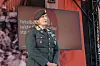 En uniformert kvinne står ved en scene med en stor skjerm og rød bakgrunn under en debatt.