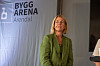 En kvinne i grønn dress står foran en scene med Bygg Arena Arendal-banner i bakgrunnen.