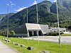 Grått industribygg ved en vei, med flaggstenger, grønne fjell og blå himmel i bakgrunnen.