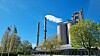 Industrifabrikk med høye skorsteiner, røyk, trær og flagg under blå himmel.