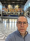 Mann med briller i en stor hall med marmorgulv og fargerik veggutsmykning.