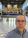 Mann med briller i en stor hall med marmorgulv og fargerik veggutsmykning.