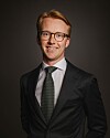 Smilende mann med briller i mørk dress og slips mot en mørk bakgrunn.