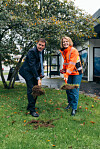 To personer med spader holder jord og planter på en gressplen foran en bygning.