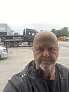 Skjeggete mann i mørk jakke foran en lastebil og anleggsmaskiner på et industriområde.