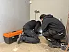 To personer kneler og arbeider med rør i et flislagt rom ved siden av en oransje verktøykasse.