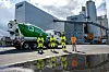 Grønn elektrisk betongbil og fire personer foran en industrifabrikk, med speilbilde i en vanndam.
