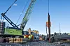Anleggsmaskiner, stålpeler og arbeidere på en byggeplass med bybebyggelse i bakgrunnen.