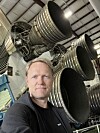 En mann tar selfie foran flere store rakettmotorer i en industrilignende hangar.