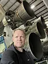 En mann tar selfie foran flere store rakettmotorer i en industrilignende hangar.