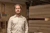 Håndverker står i et verksted med trevirke og verktøy i bakgrunnen.