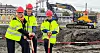 Tre personer i verneutstyr står ved en byggeplass med gravemaskiner i bakgrunnen.
