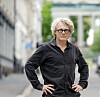 En gråhåret person med briller og mørk skjorte står med hendene i siden i en bygate.