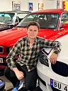 En mann sitter mellom en rød klassisk BMW og en hvit BMW i en garasje.